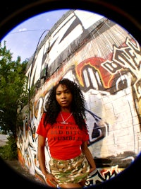 a woman wearing a camouflage shirt standing in front of a graffiti covered wall